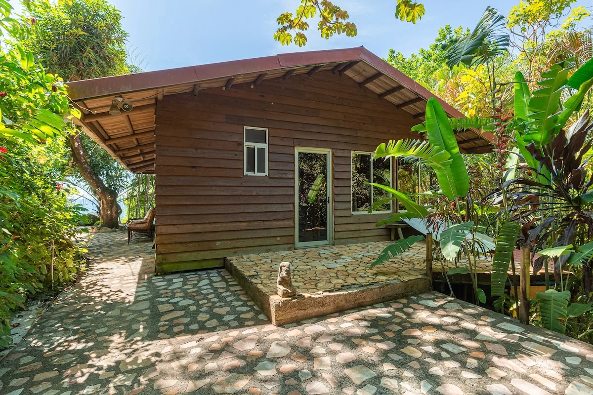 Una casa de madera está rodeada de plantas tropicales junto a un camino de piedra.