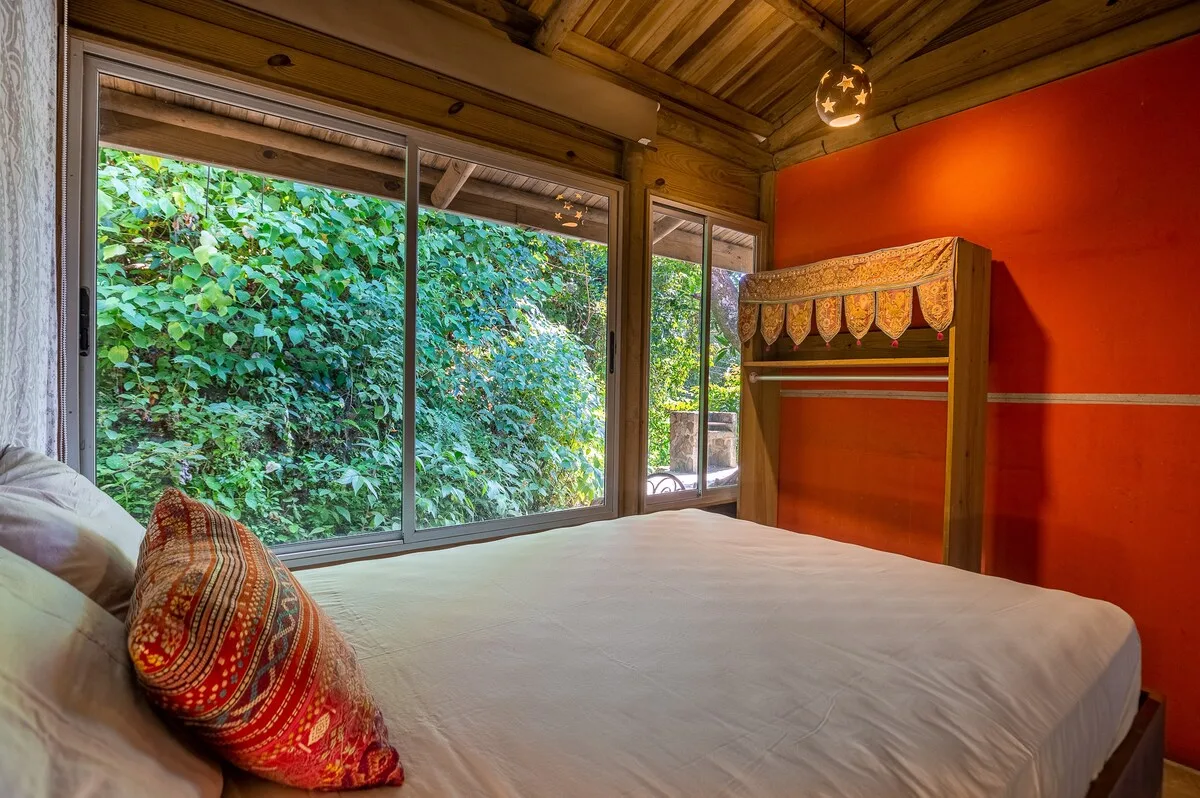 Dormitorio con una cama junto a grandes ventanas que dan a una vegetación frondosa.