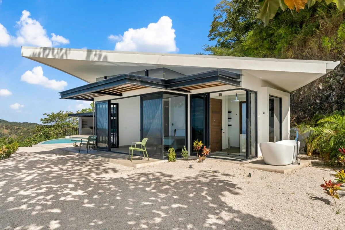 A modern house with broad roof overhangs and glass doors opens onto a sunny patio.