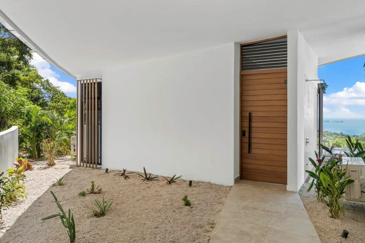 Exterior entryway with a wooden door, sandy landscaping, and a glimpse of the sea.