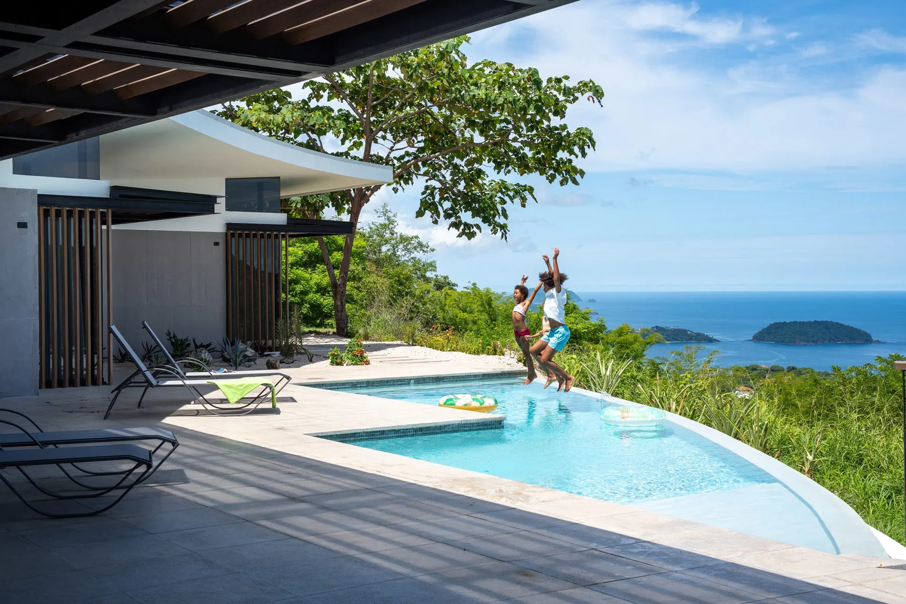 An outdoor pool beside a shaded patio overlooks green hills and the sea.