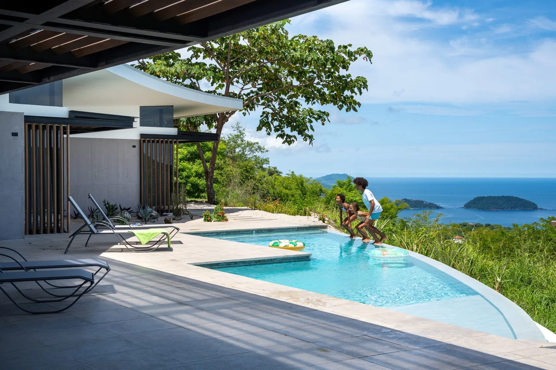 An outdoor pool beside a deck overlooks the ocean and a green island.