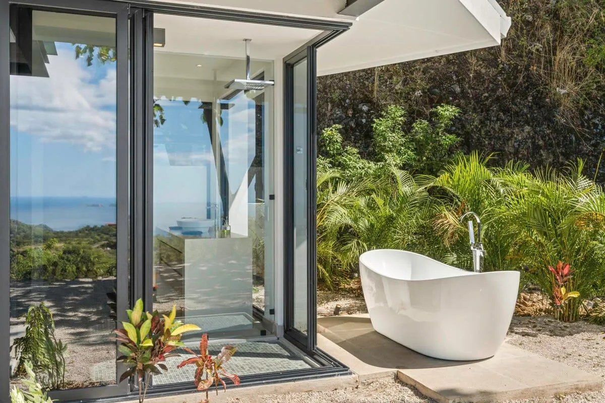 An outdoor bathtub beside an open glass doorway, surrounded by tropical plants.