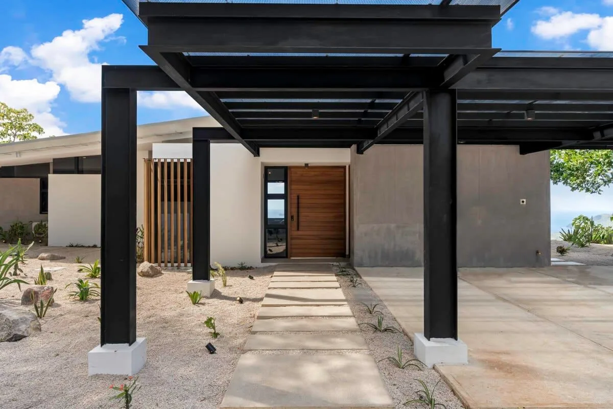 A modern house entrance with a wooden front door beneath a dark pergola.