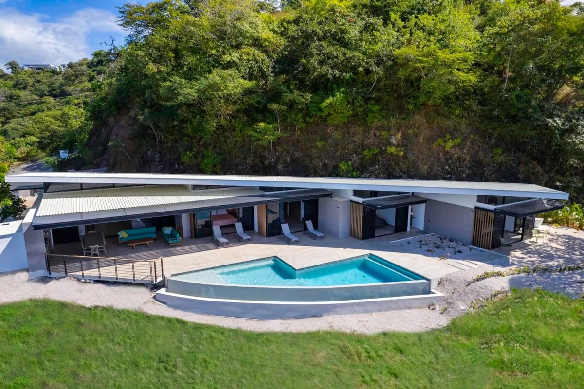 A modern outdoor pool sits in front of a low building surrounded by greenery.