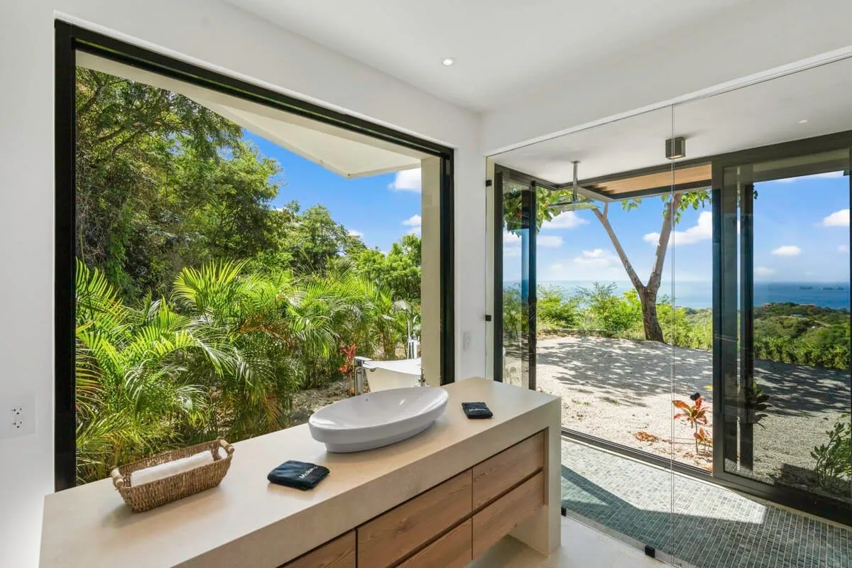 A bright bathroom vanity overlooks lush greenery and a view of the sea.