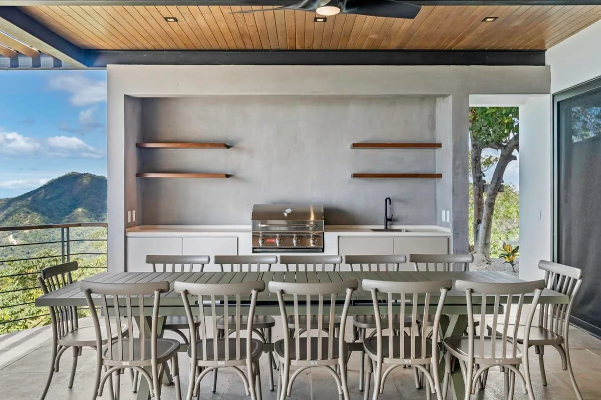 An outdoor dining area with a long table and built-in grill overlooks a mountain landscape.
