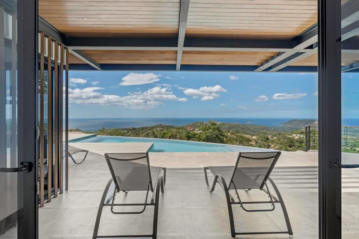 An open terrace overlooks a swimming pool and the sea, with two lounge chairs in front.