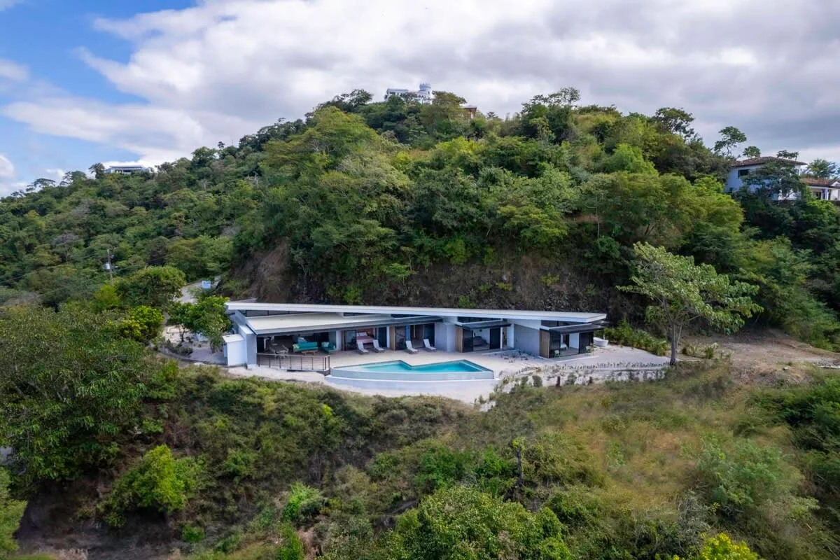 A modern low-rise building with a swimming pool sits among dense green hills.