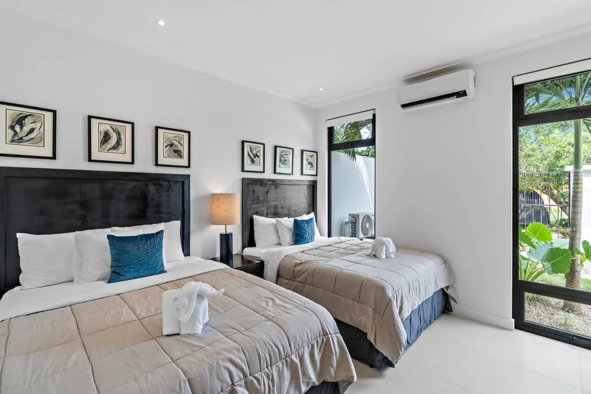 A bright bedroom with two beds, framed artwork, and large windows.