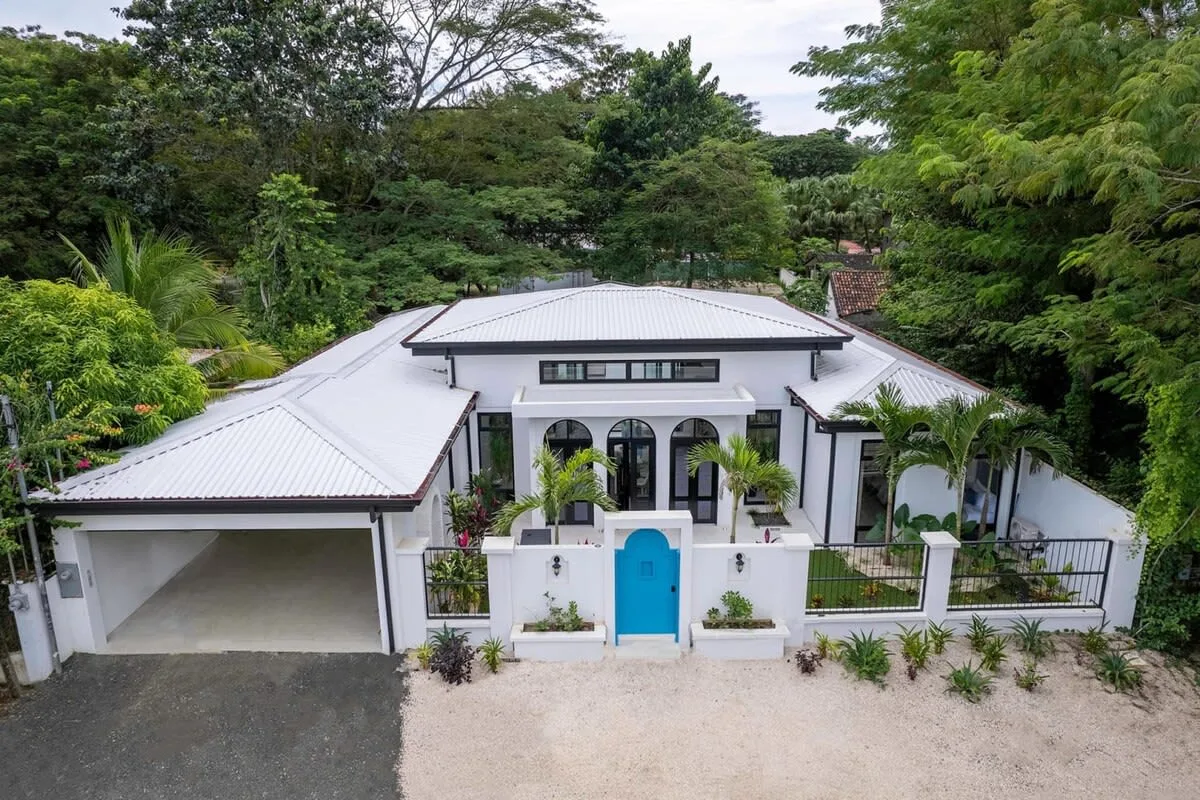 A white house with a bright blue front door is surrounded by lush greenery.