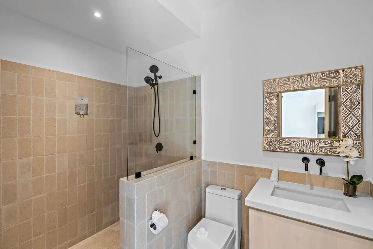 A tiled bathroom with a glass shower, toilet, and sink beneath a decorative mirror.