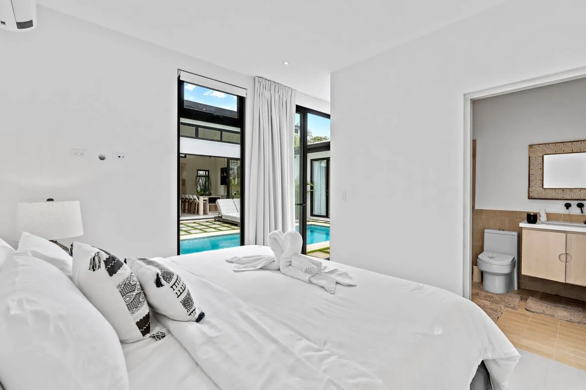 A bright bedroom with white bedding, large windows, and an adjoining bathroom.