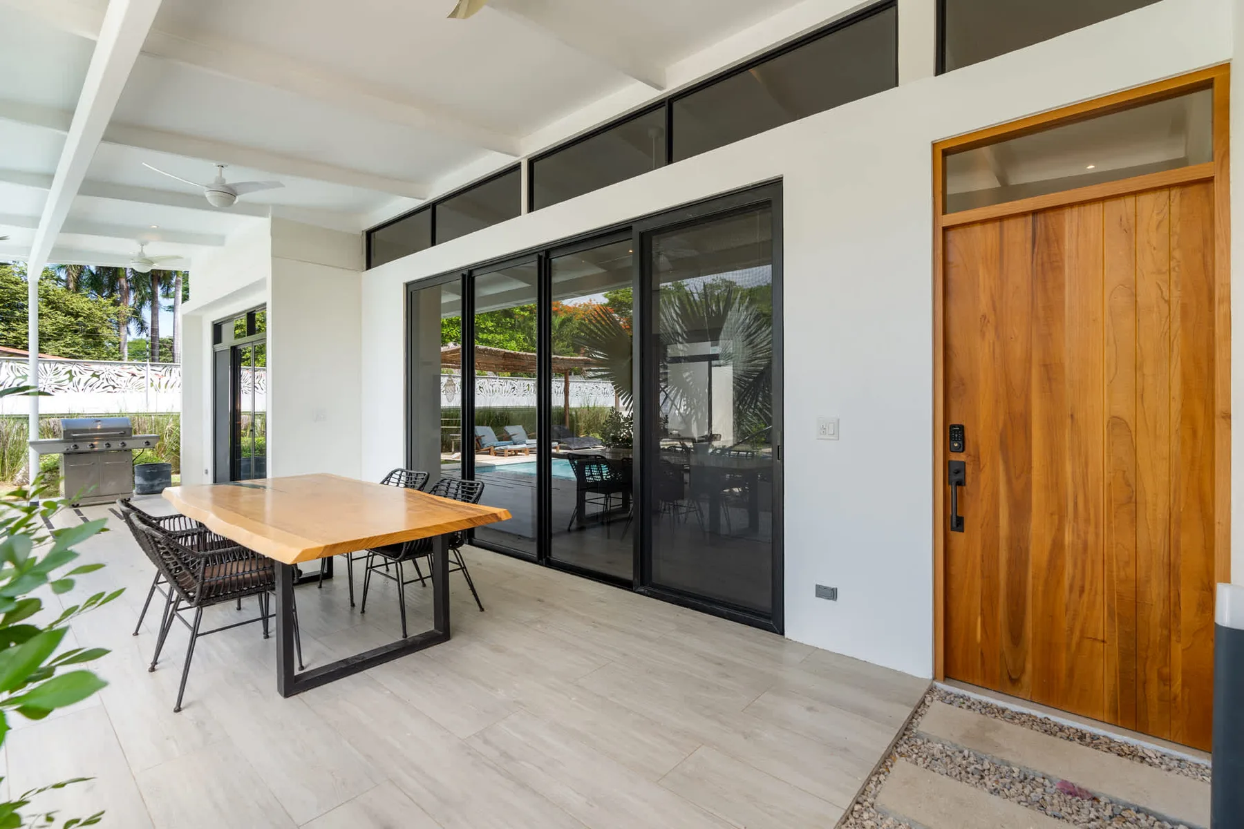 A covered patio has a dining table beside large glass doors and a wooden entrance door.