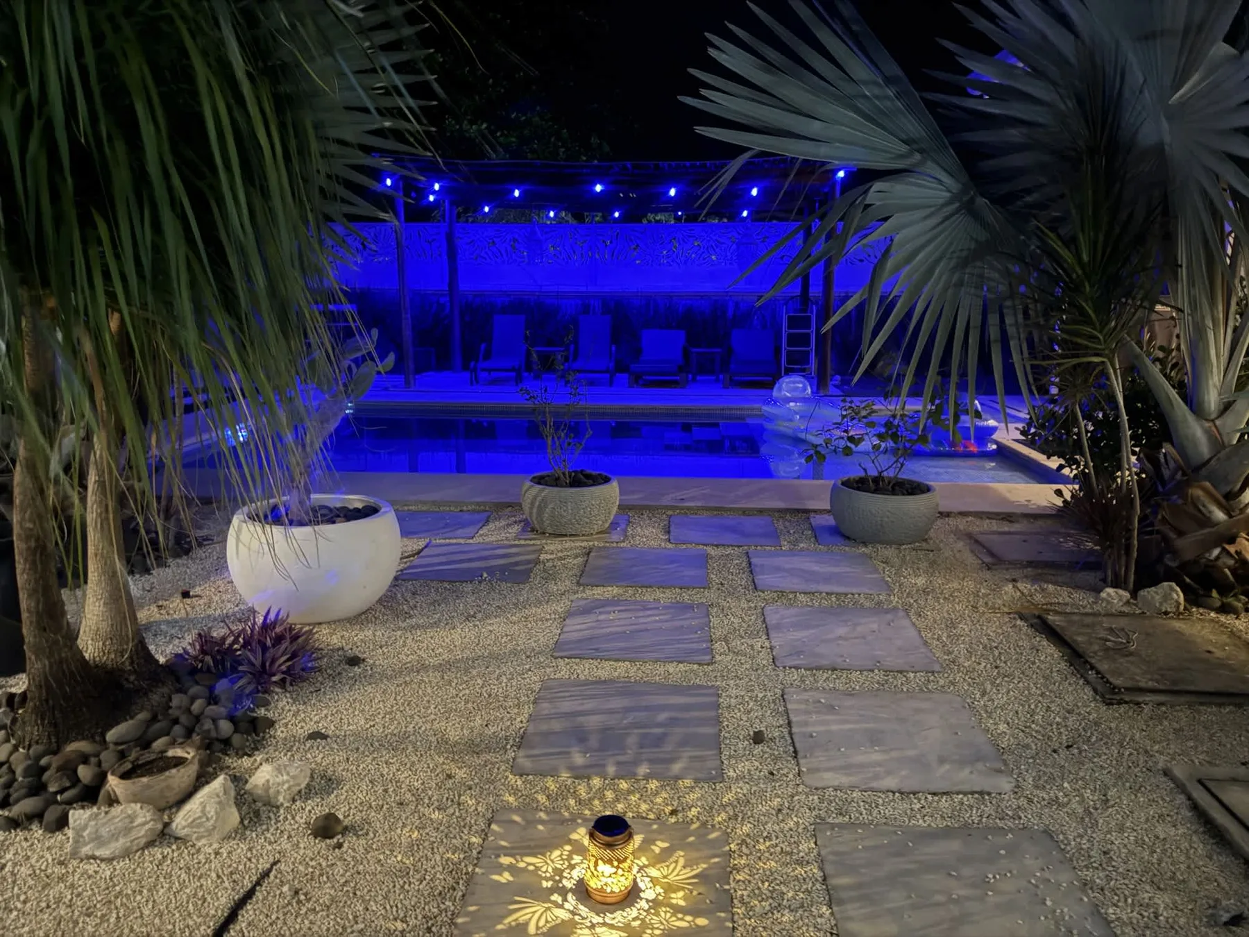 A nighttime pool area glows blue beyond a gravel path lined with stepping stones.