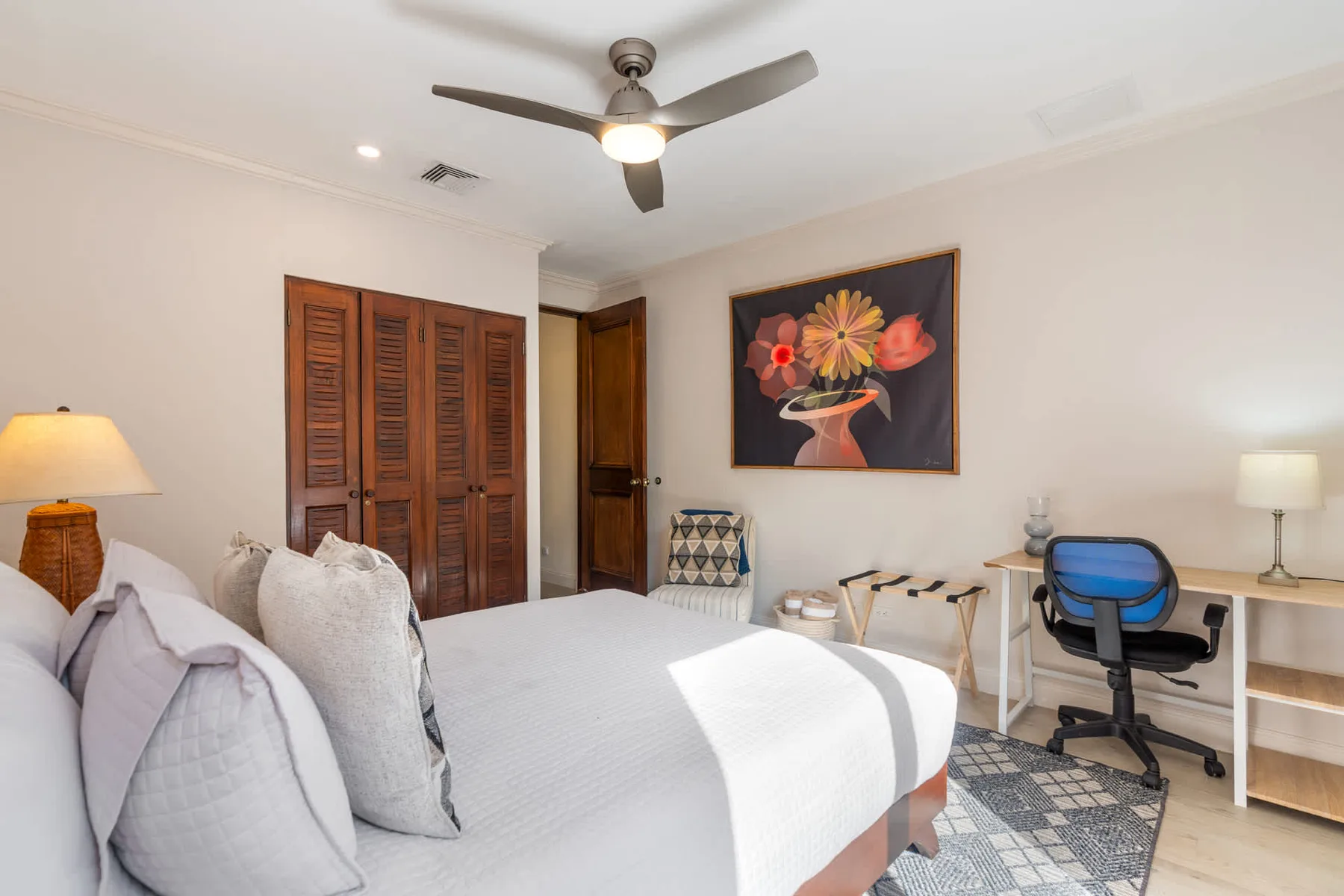 Un dormitorio luminoso con una cama bien arreglada y un escritorio bajo un cuadro floral.