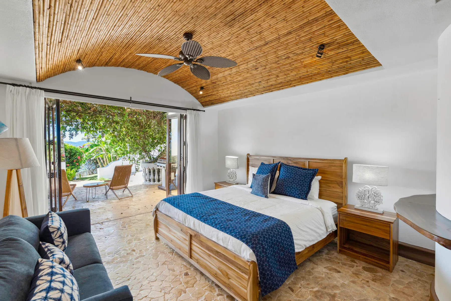 A bedroom with a wooden bed and blue bedding opens onto a leafy patio.