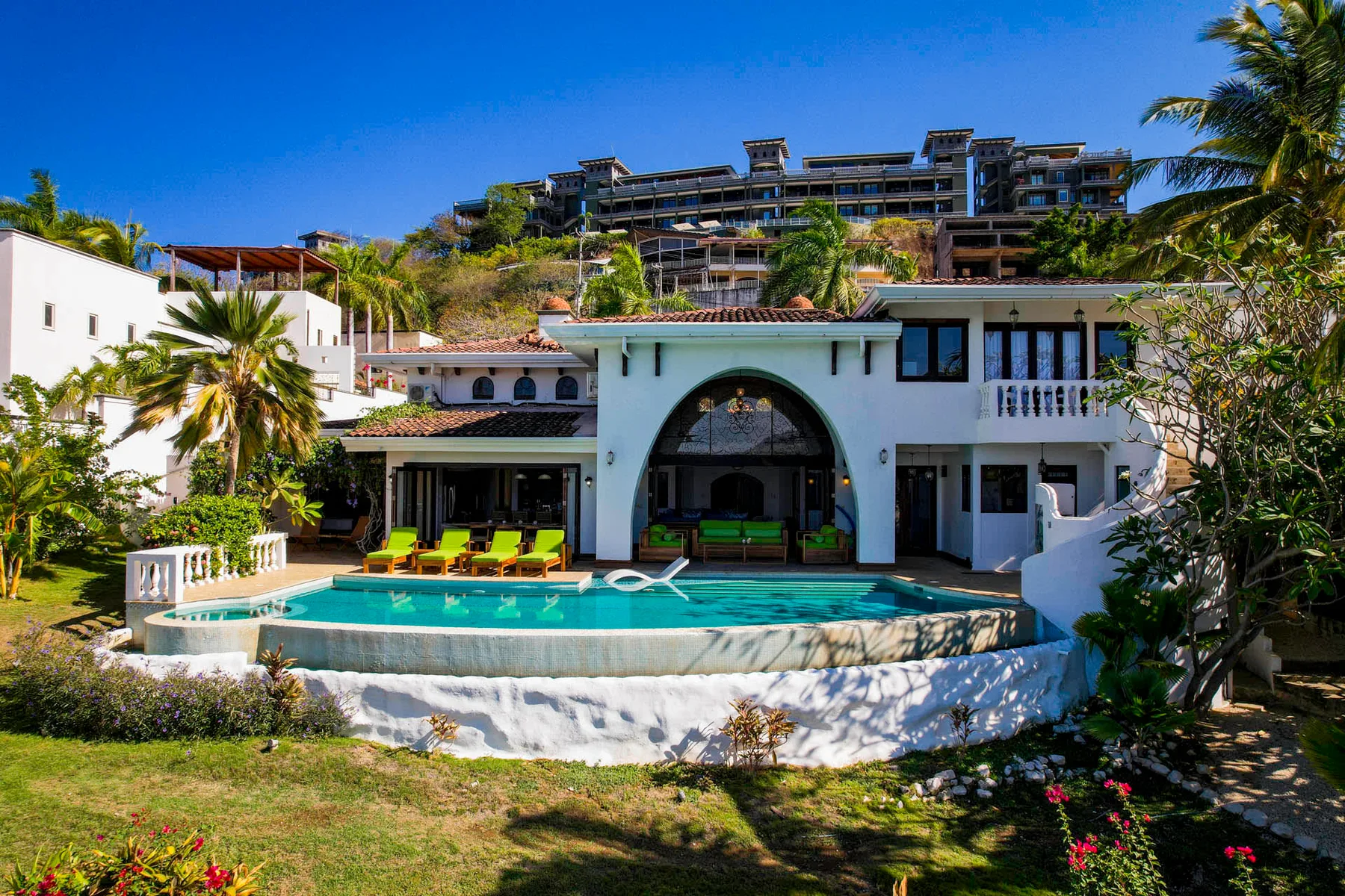A white villa with an arched entrance overlooks a curved swimming pool and garden.