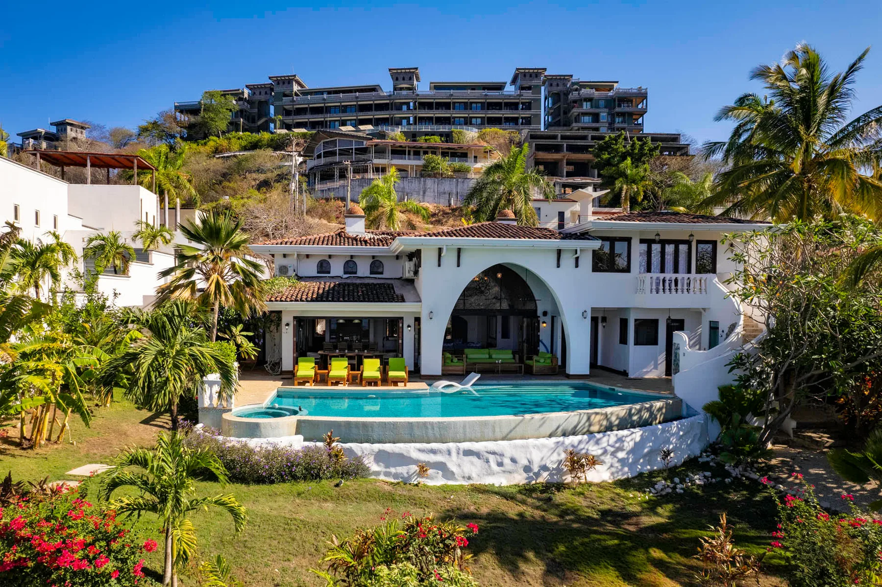A white villa with an arched entrance overlooks a curved swimming pool and landscaped garden.