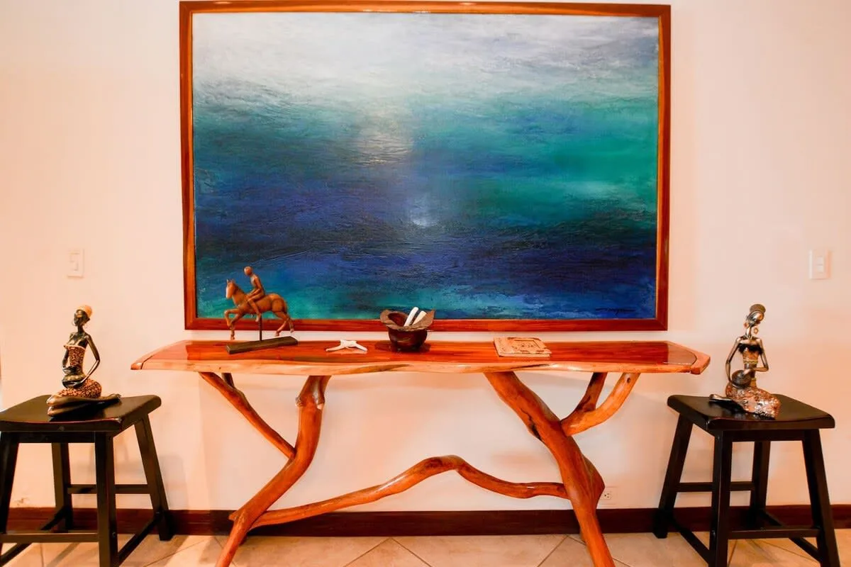 A large seascape painting hangs above a sculptural wooden table, with small statues on either side.