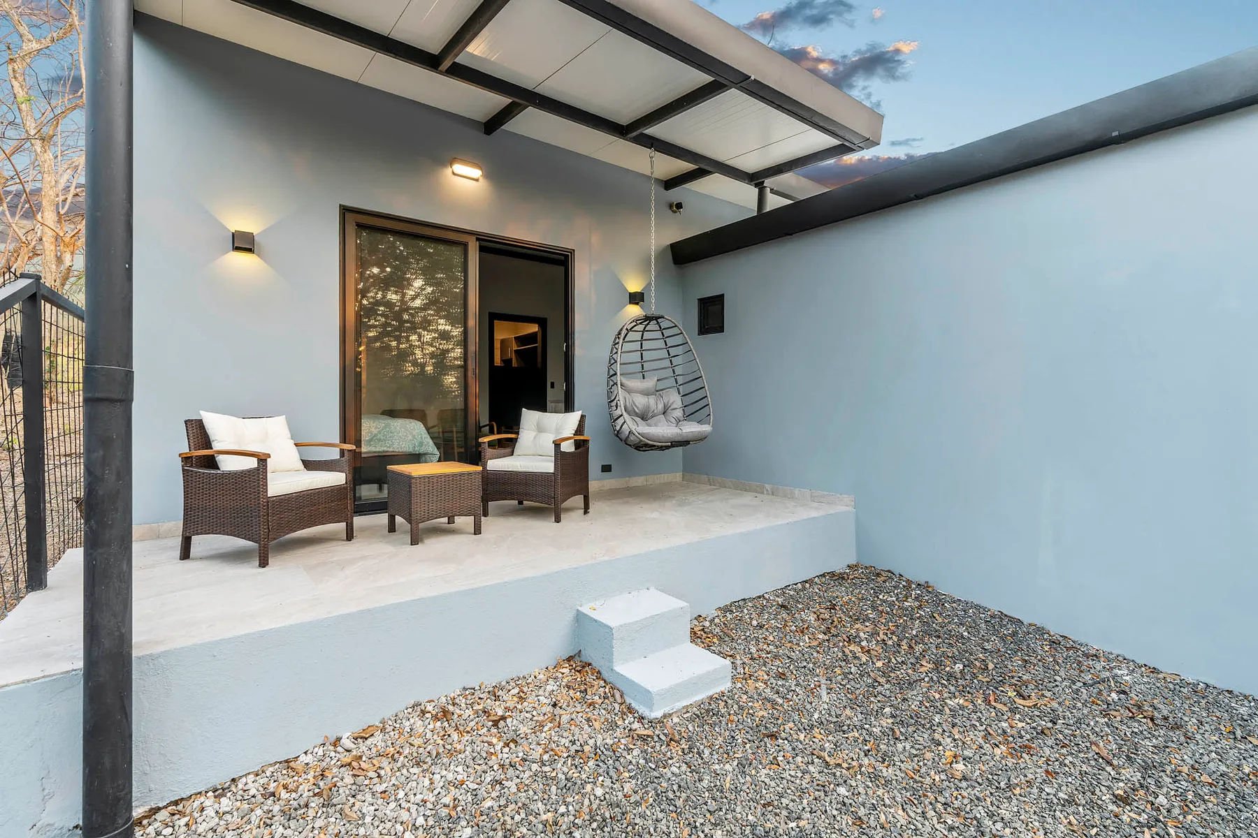 A covered outdoor patio has seating and a hanging chair beside a gravel yard.