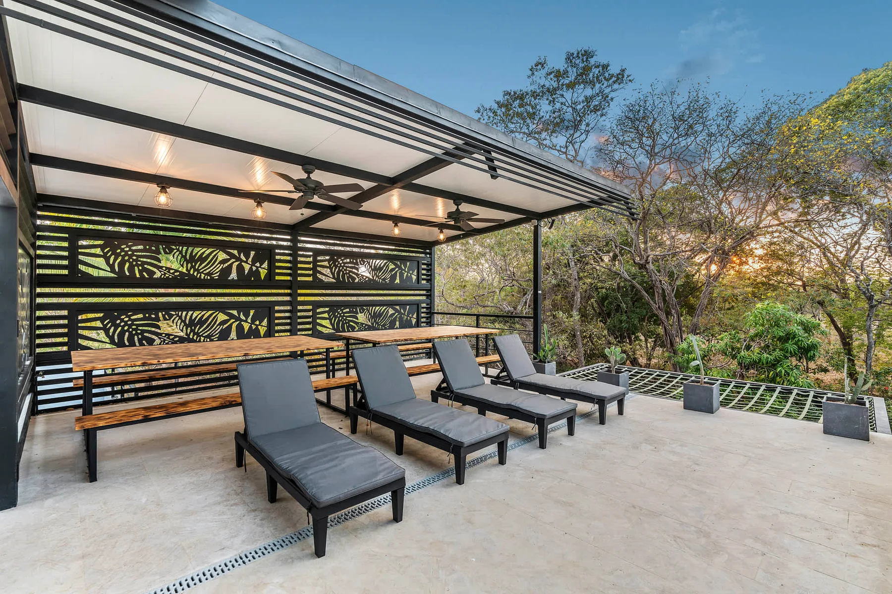 A covered outdoor patio has lounge seating, a long table, and leafy surroundings.