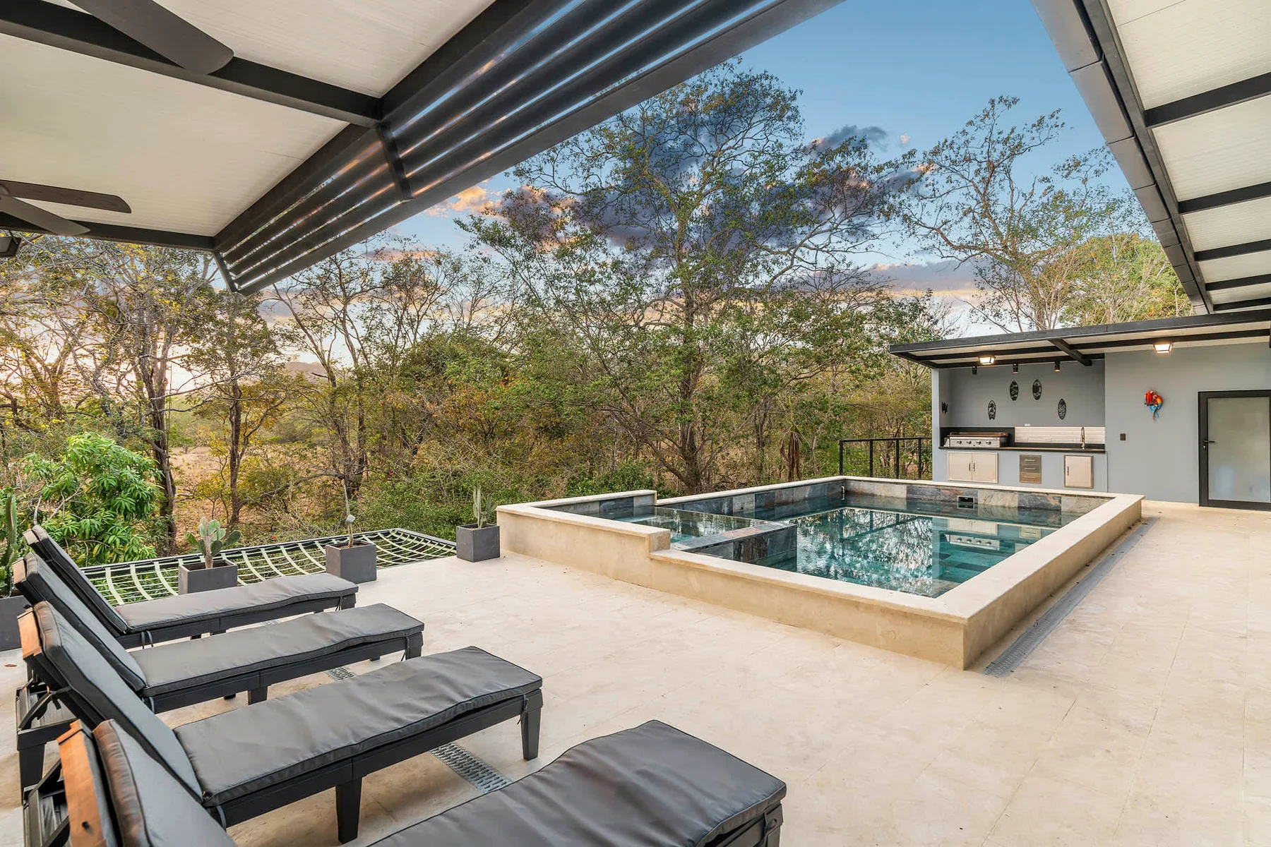 A covered outdoor patio with a pool and lounge seating overlooks trees.