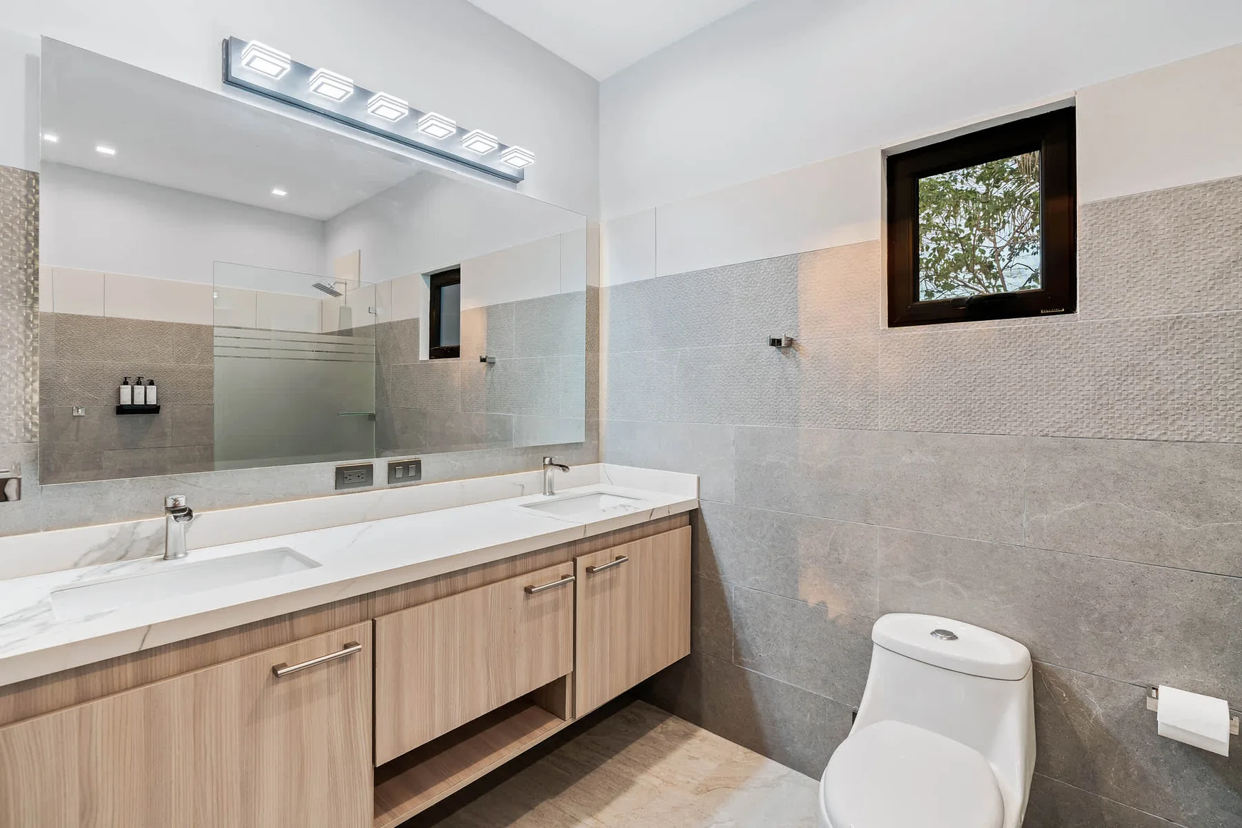 A bright bathroom with a long vanity, large mirror, and window.