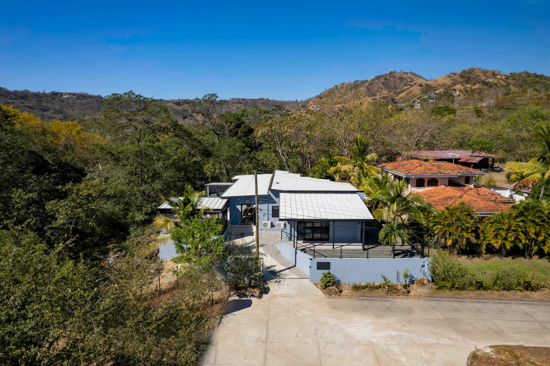 A house sits among trees and palms, with hills rising in the background.