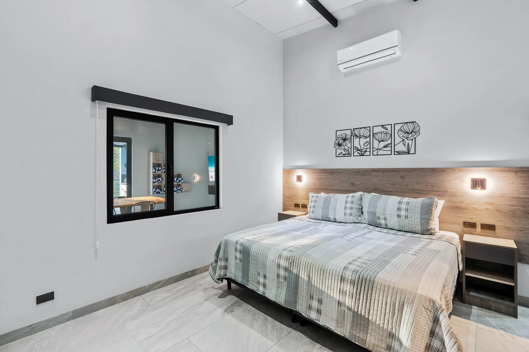 A bedroom with a neatly made bed and a window into an adjoining room.