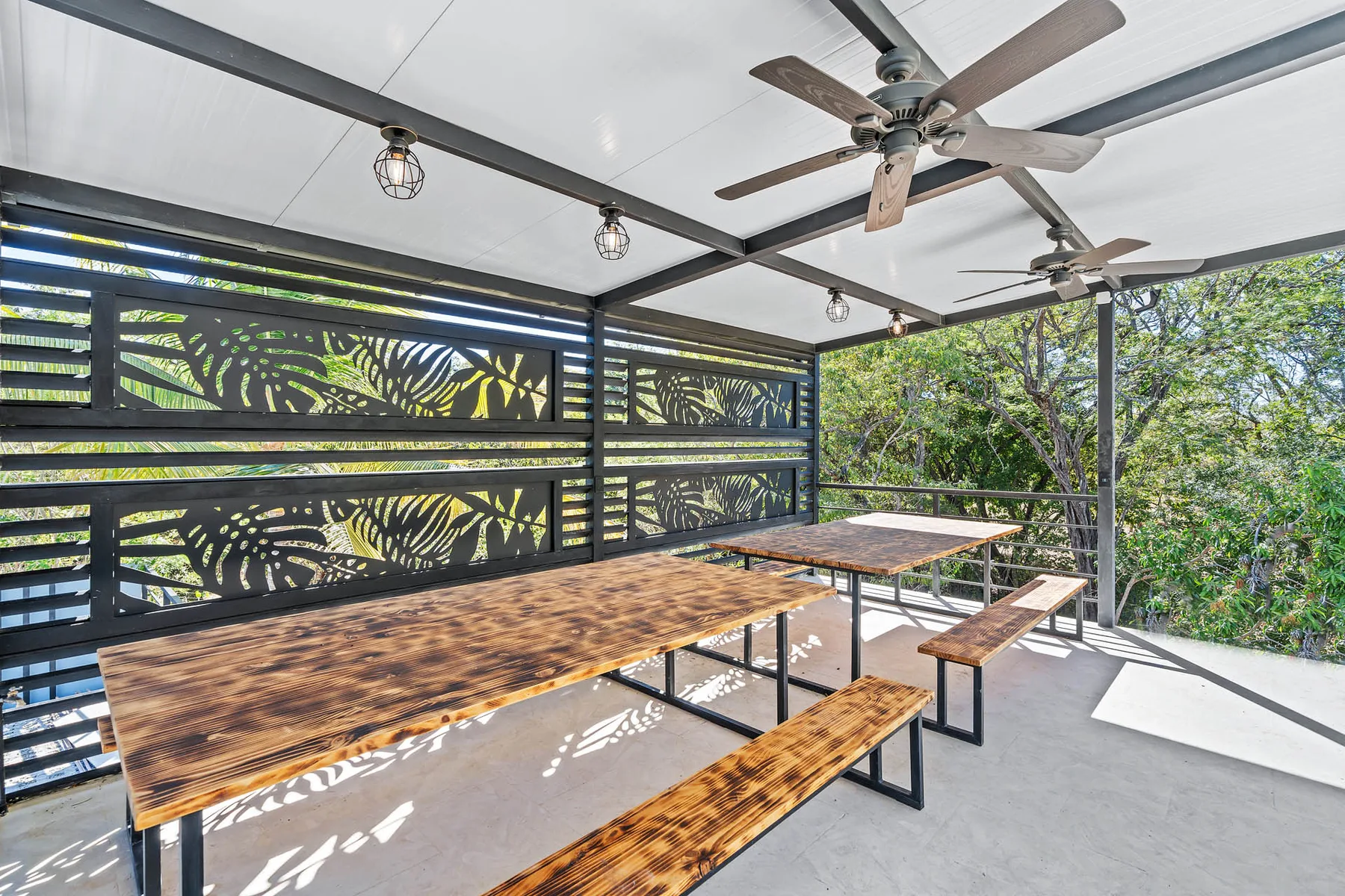 A covered outdoor dining area has long wooden tables and benches beneath ceiling fans.