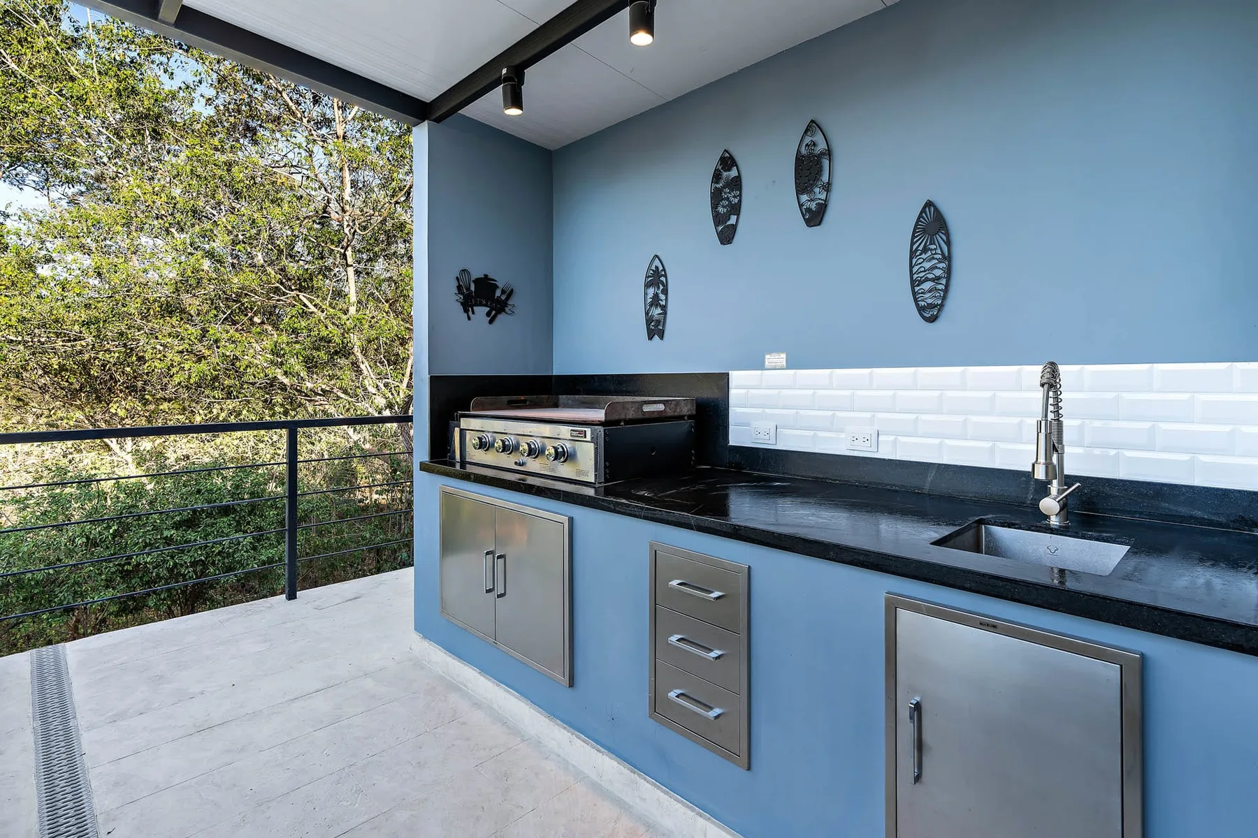 An outdoor kitchen with a countertop sink and trees visible beyond the railing.