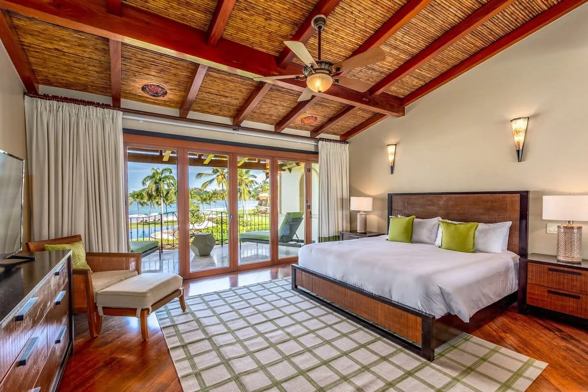A bedroom with a large bed and sliding glass doors opening onto a palm-lined view.