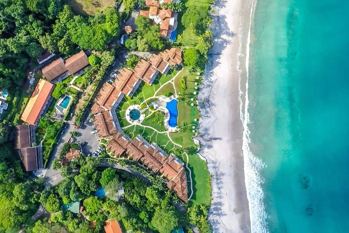 Shared amenity: Aerial view of a beachfront resort surrounded by greenery, with pools and terracotta-roofed buildings.