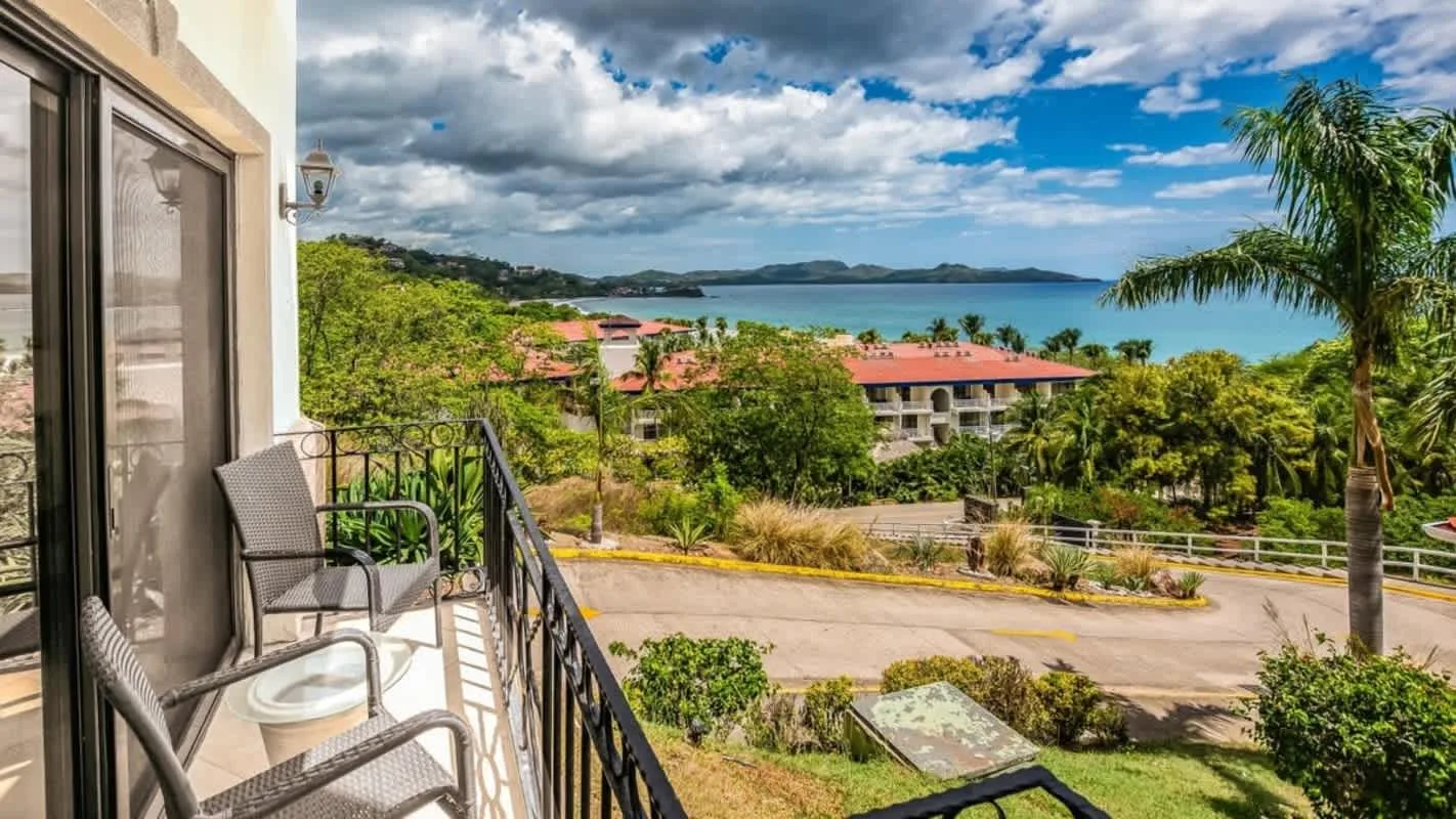 A balcony overlooks a landscaped road, palm trees, and a bay beneath a cloudy sky.