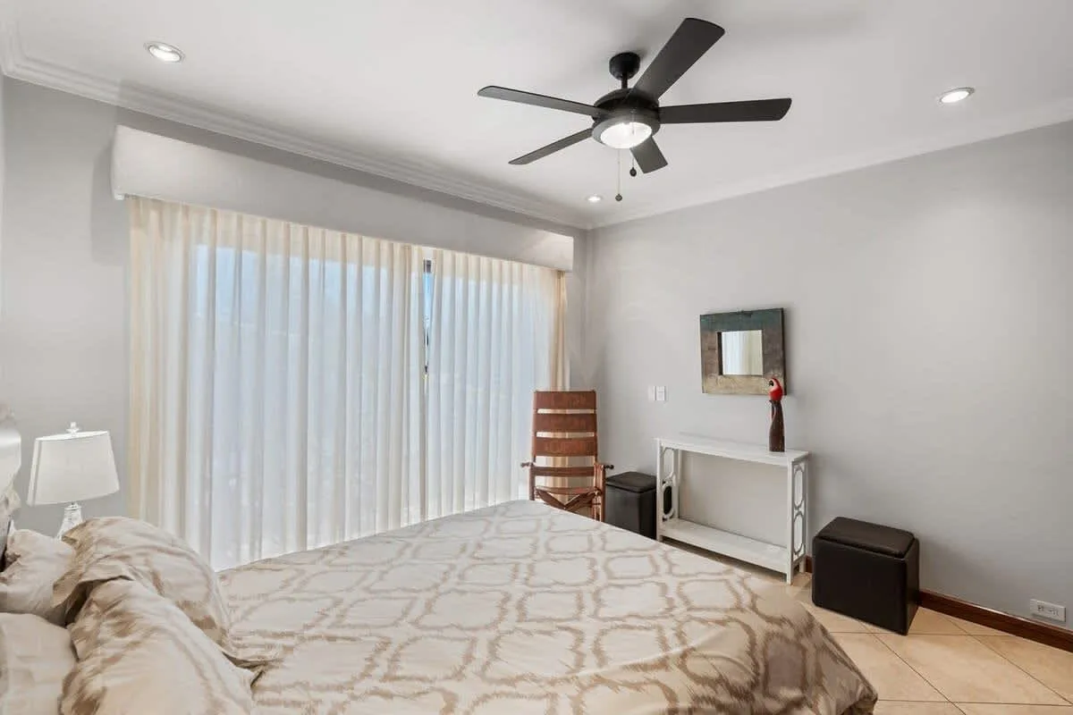 A bright bedroom with a bed, sheer curtains, and a ceiling fan.