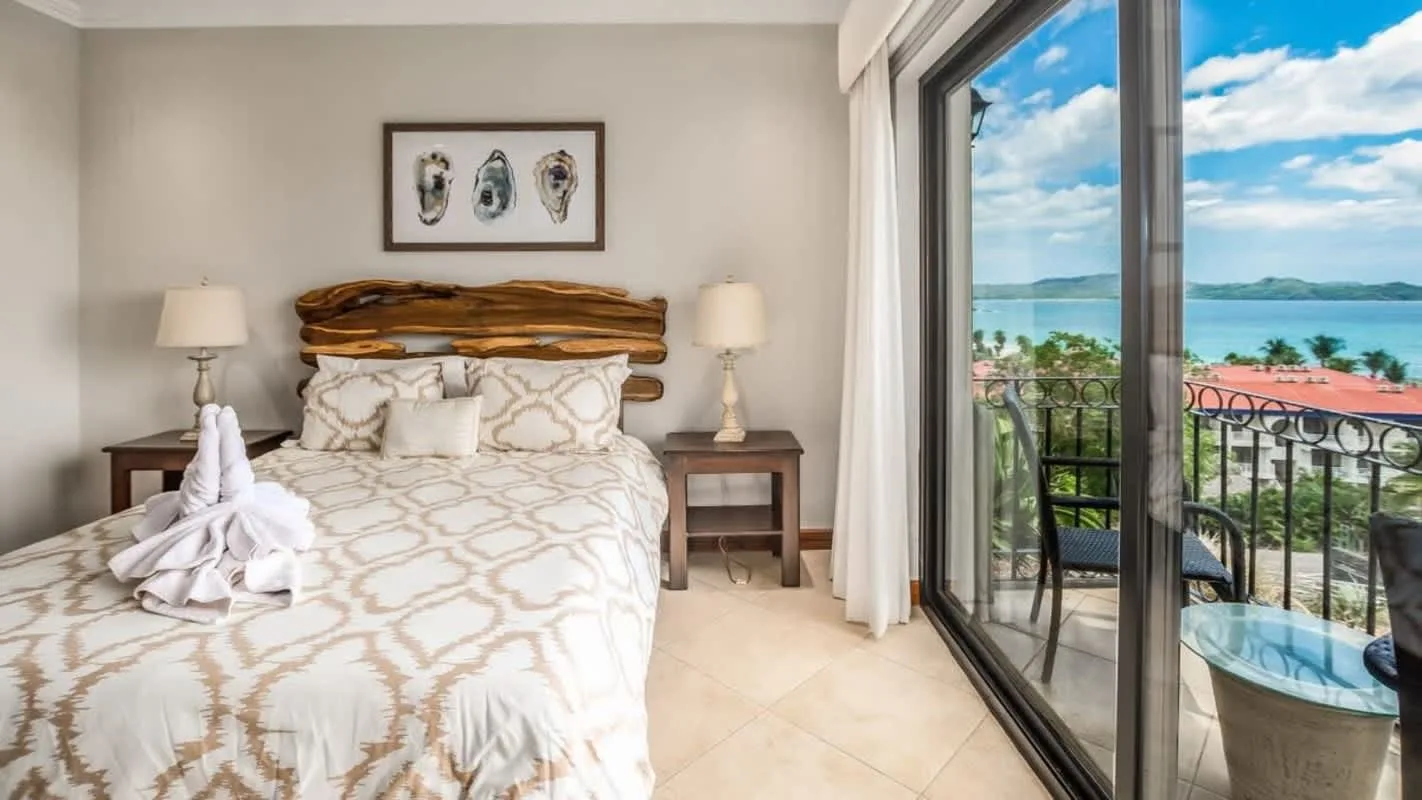 A bedroom with a bed beside glass doors opening onto a balcony with a sea view.