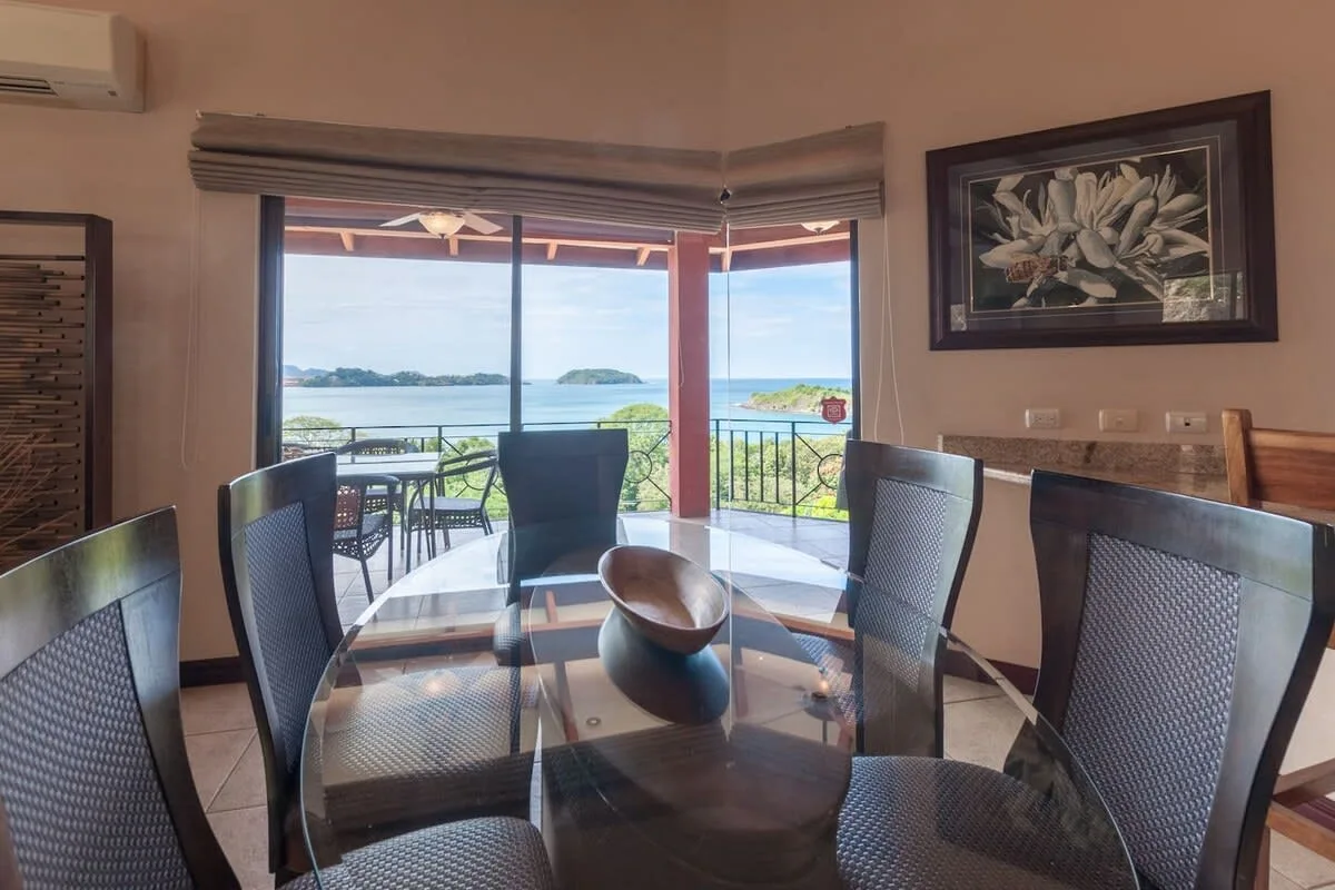 A dining area with a glass table and chairs overlooks the water through open windows.