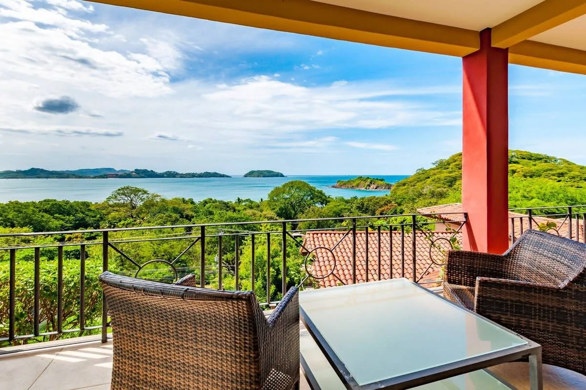 A covered terrace has seating and a glass-topped table overlooking the sea and green hills.