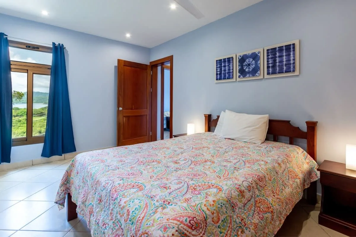 A bedroom with a patterned bedspread and a window framed by blue curtains.