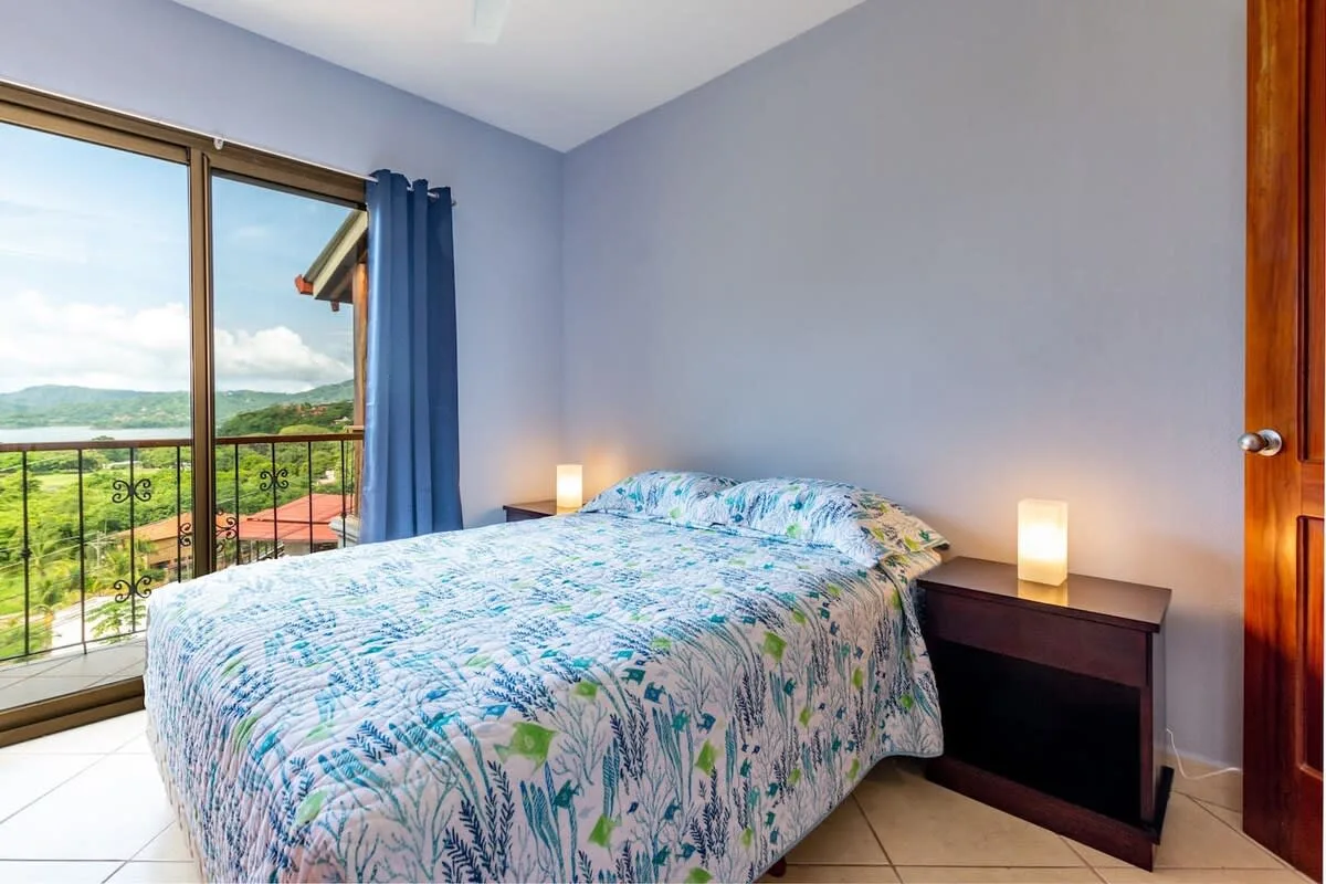 A bedroom with a bed beside a large window overlooking green hills.