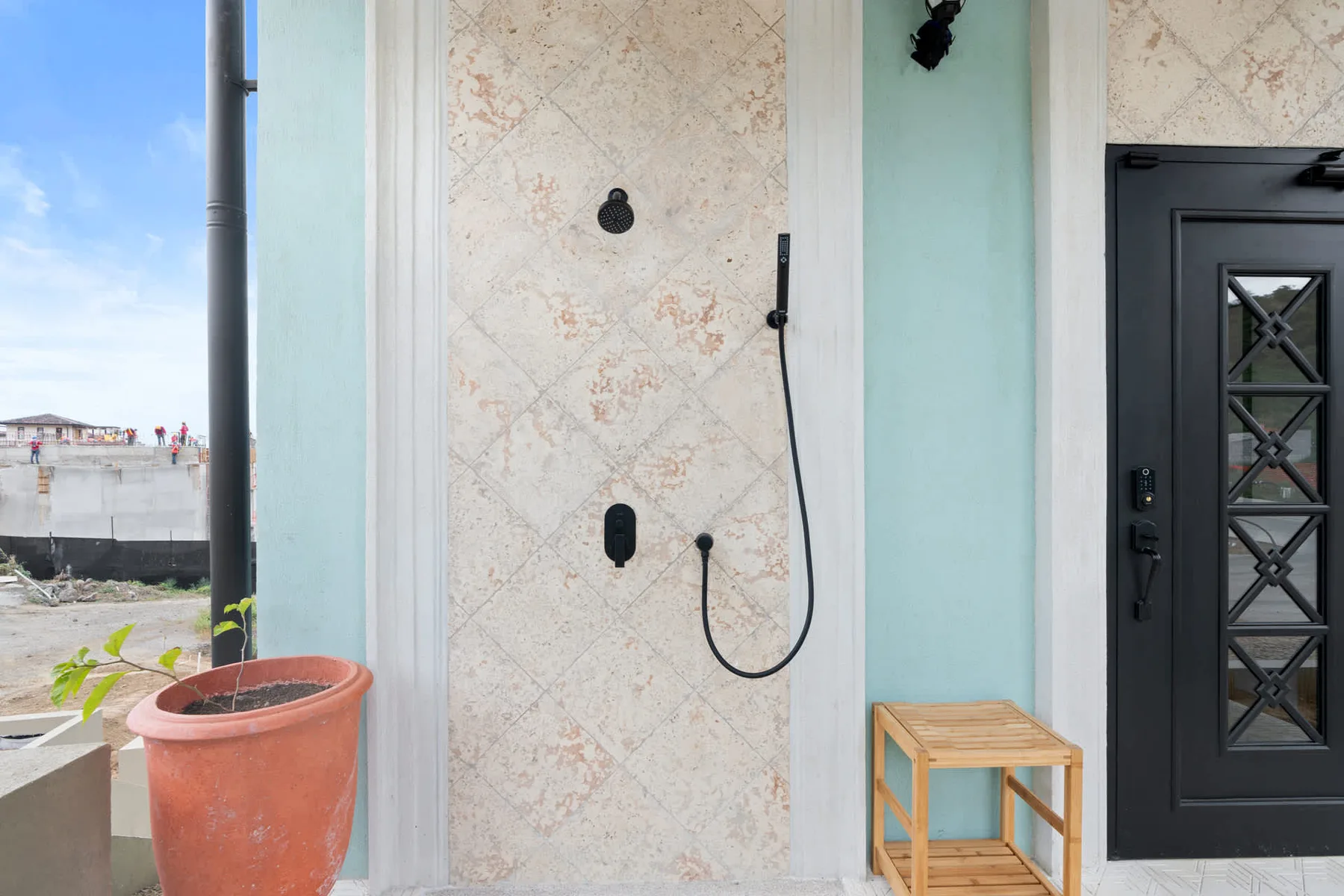 Una zona de ducha exterior junto a una puerta oscura y una planta en maceta.