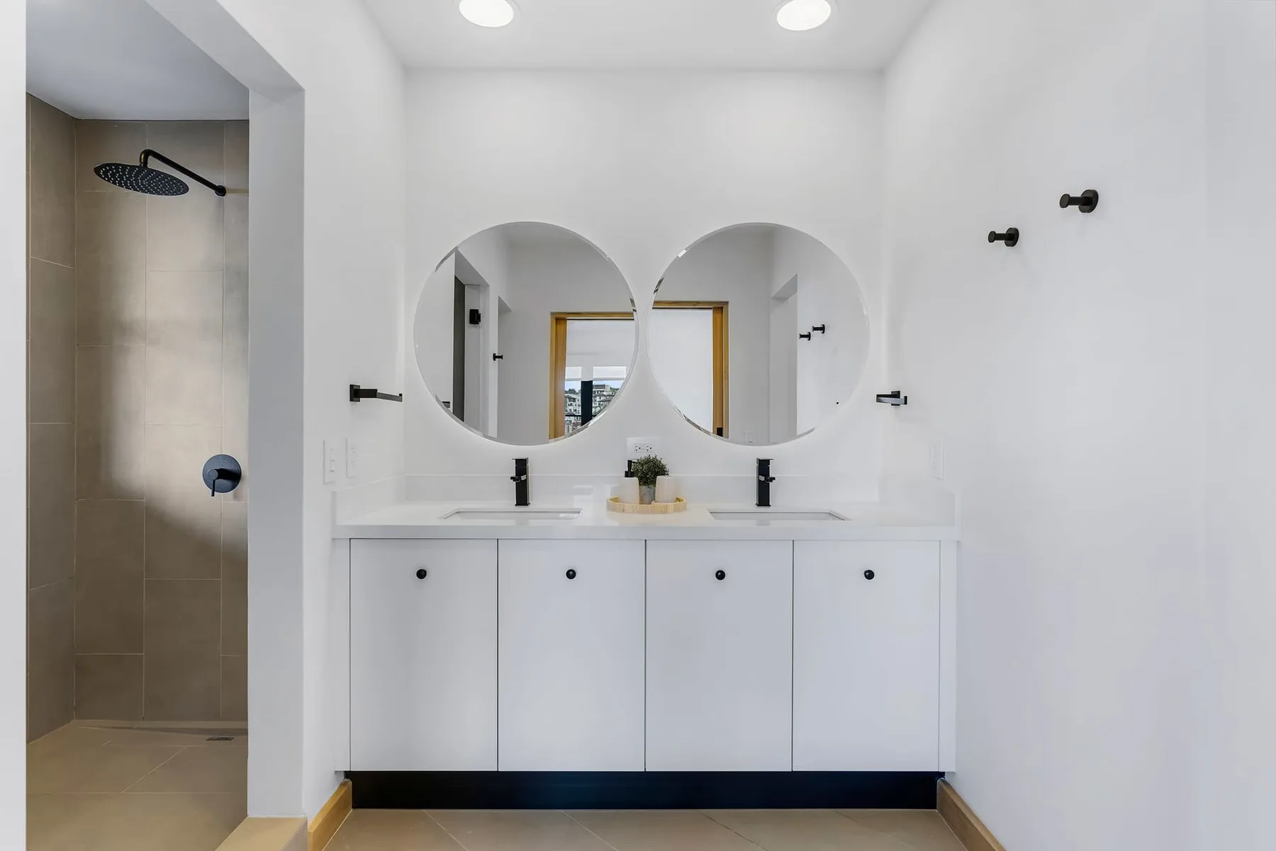 Un baño luminoso tiene un tocador doble, espejos redondos y una ducha abierta.
