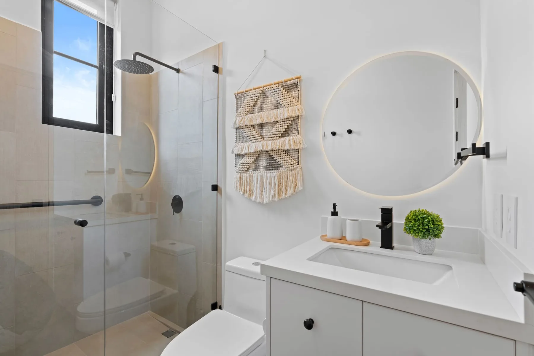 Un baño luminoso con una ducha de vidrio y un tocador blanco bajo un espejo redondo.