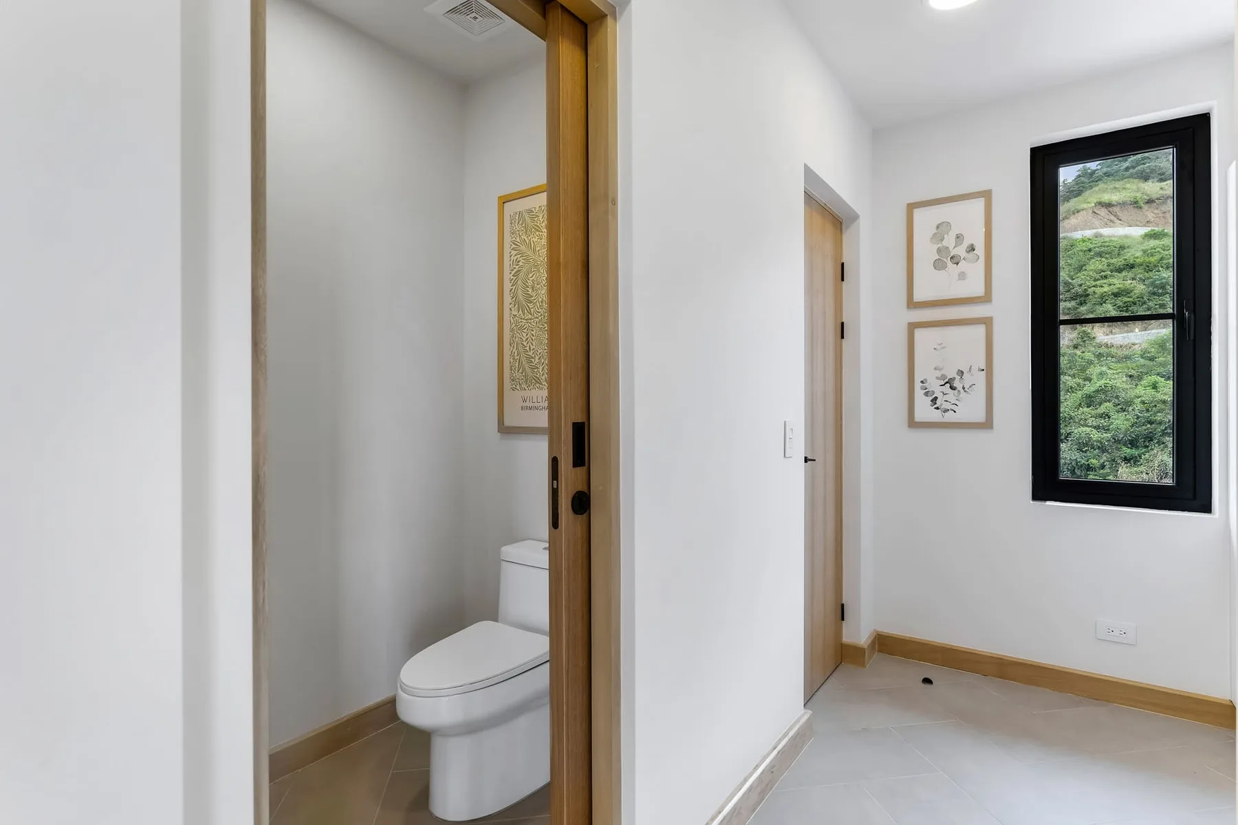 Un pasillo luminoso con una ventana que da a colinas verdes y una puerta de baño abierta.