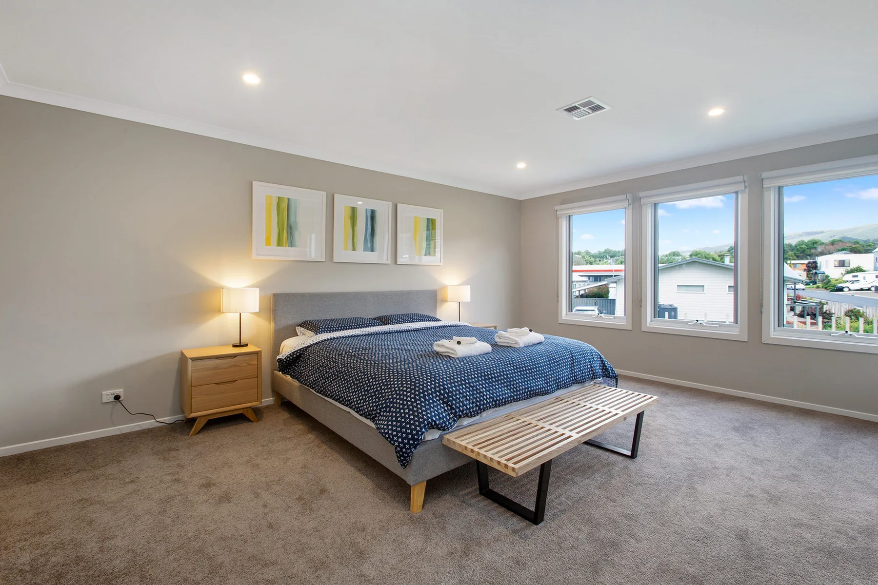 A bedroom with a neatly made bed and large windows overlooking nearby buildings.