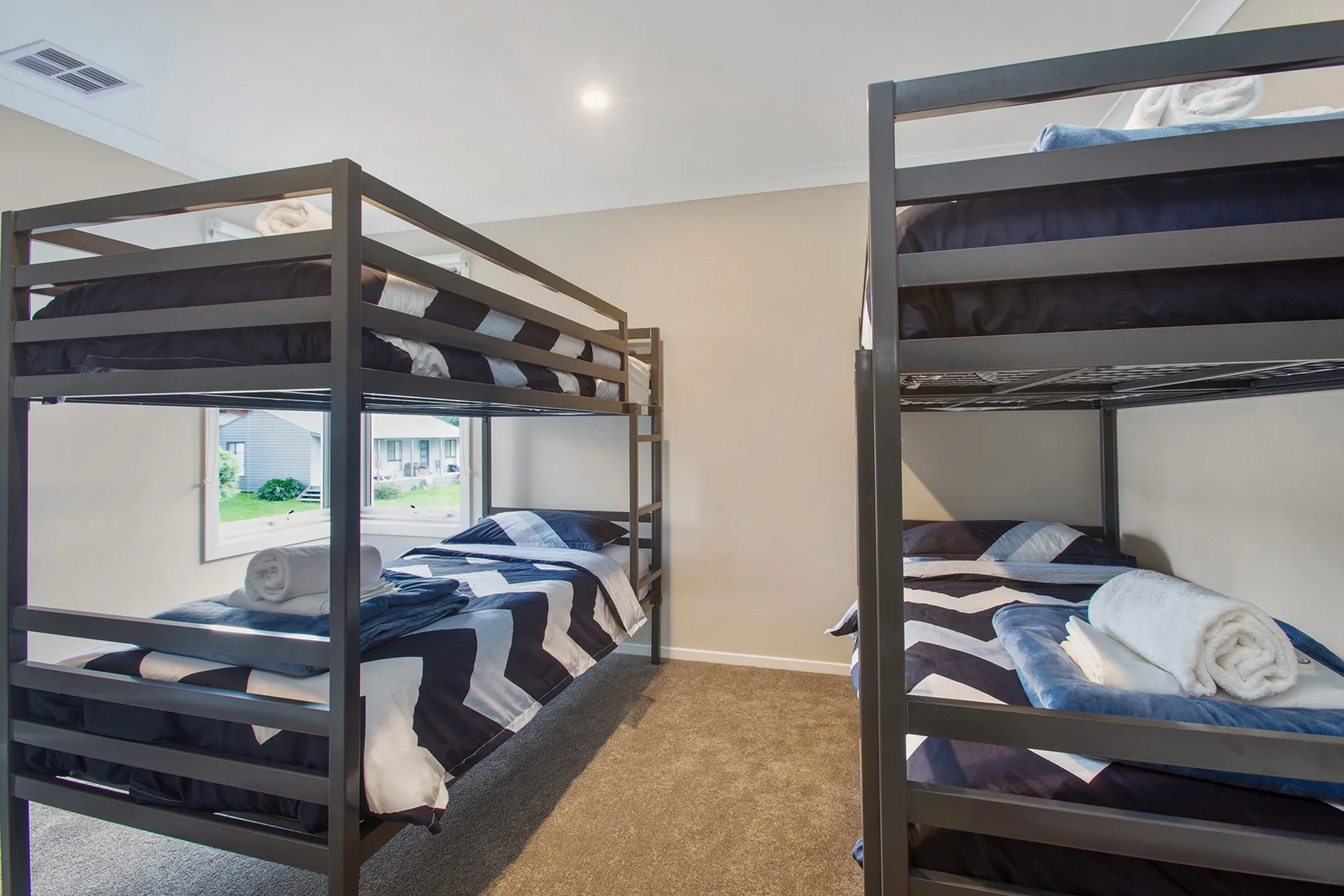 A bunk bedroom with dark bedding and a window overlooking nearby houses.