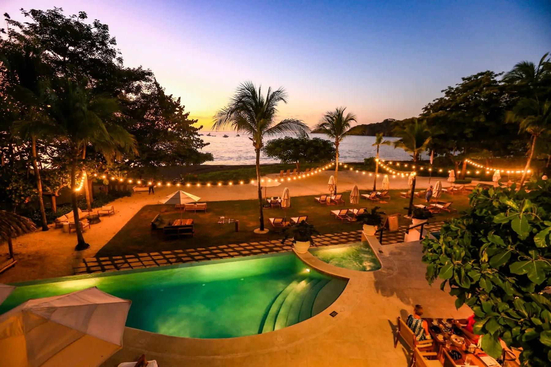 Zona común: Un patio junto a la piscina, con asientos y luces, da a la playa al atardecer.