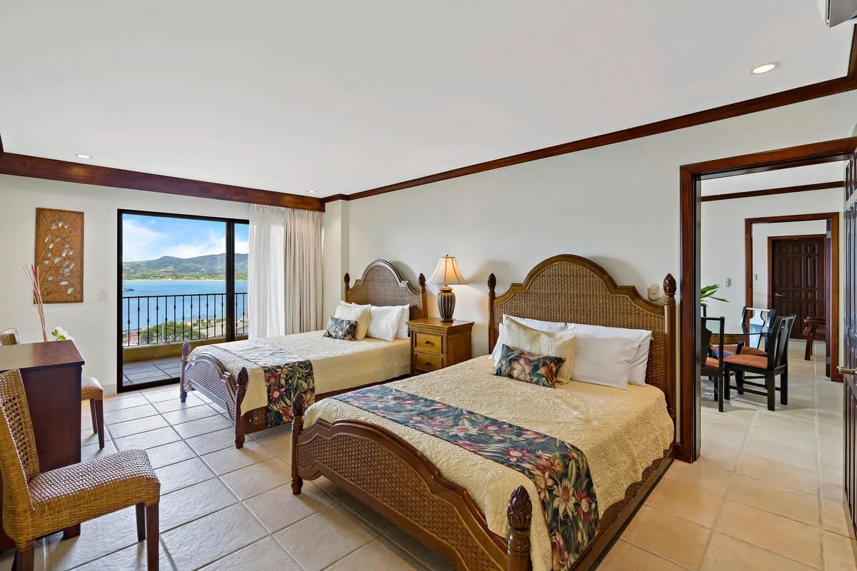 A bedroom with two beds, bedside tables, and a sliding glass door opening to a balcony.