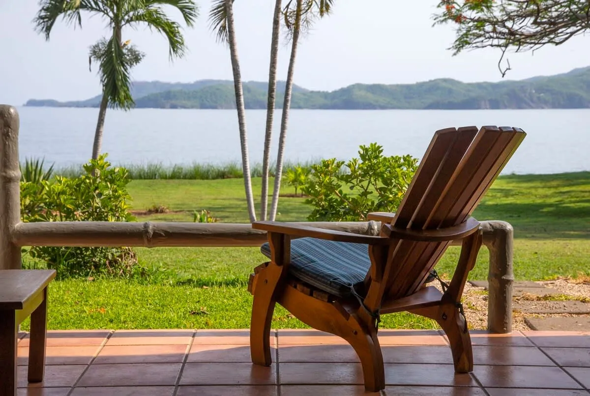 A wooden lounge chair sits on a tiled patio overlooking a lake and greenery.